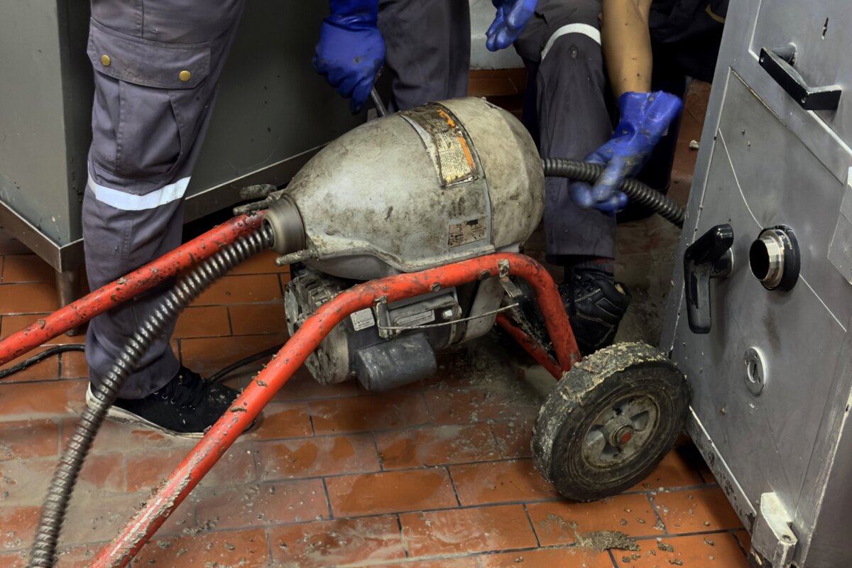 A drain cleaning machine on a tiled floor with its cable running out of the drum, gloved hands working at it and debris on the floor