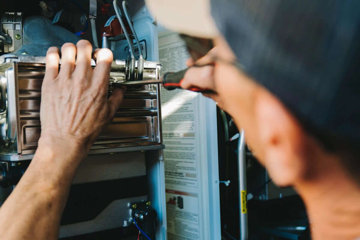 A plumber's hands and a tool inside the open cabinet of a wall-hung water heater
