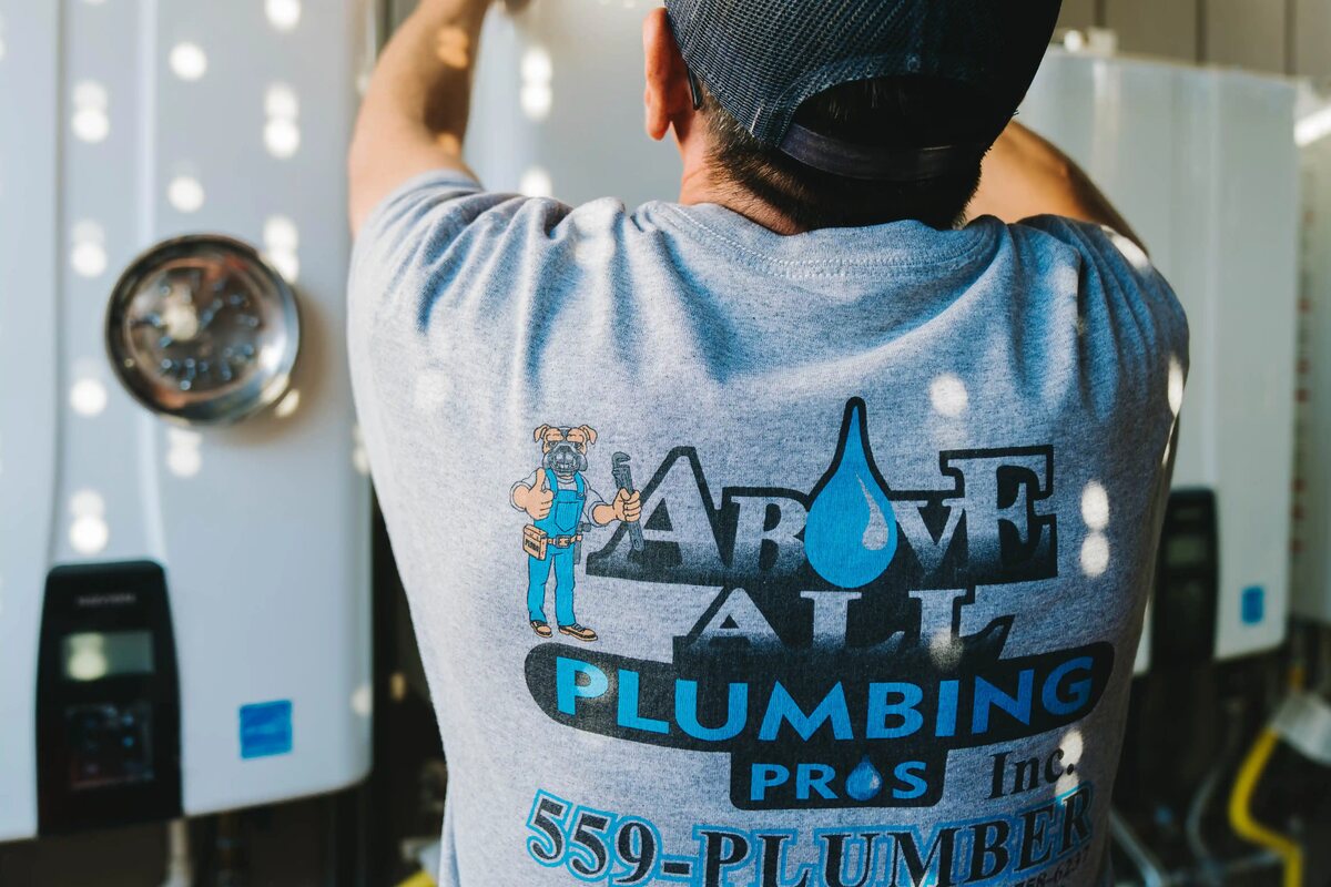 A technician, seen from behind in an Above All Plumbing shirt, working at a wall-hung water heater