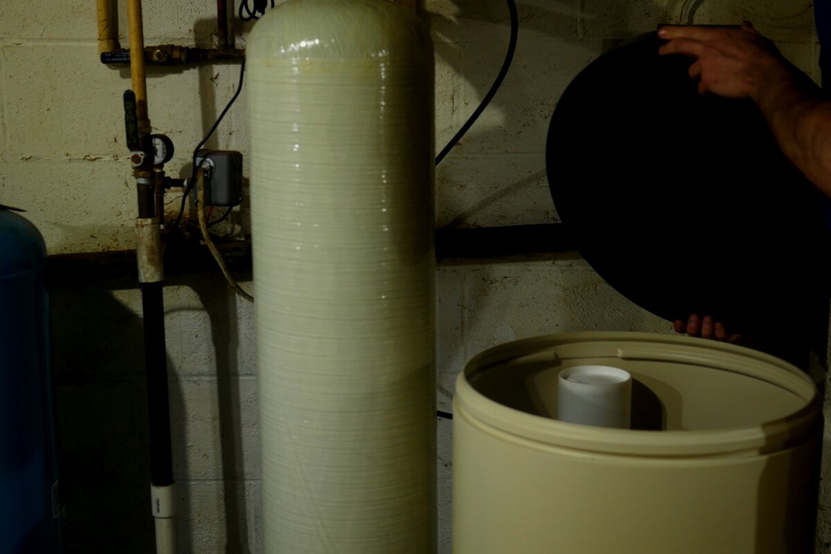 A tall water treatment tank with its brine tank and a blue pressure tank beside it, piped in a utility area